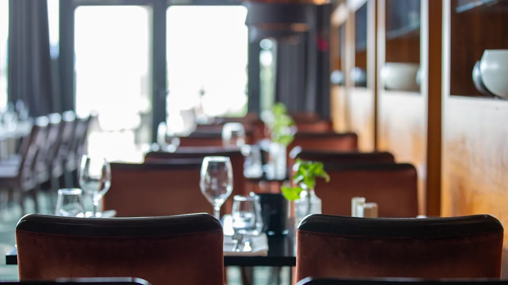 Bord dekket med glass og tallerkener venter på gjester i en mykt opplyst restaurant, med brune stoler og dekorative planter, med store vinduer som gir naturlig lys i bakgrunnen.