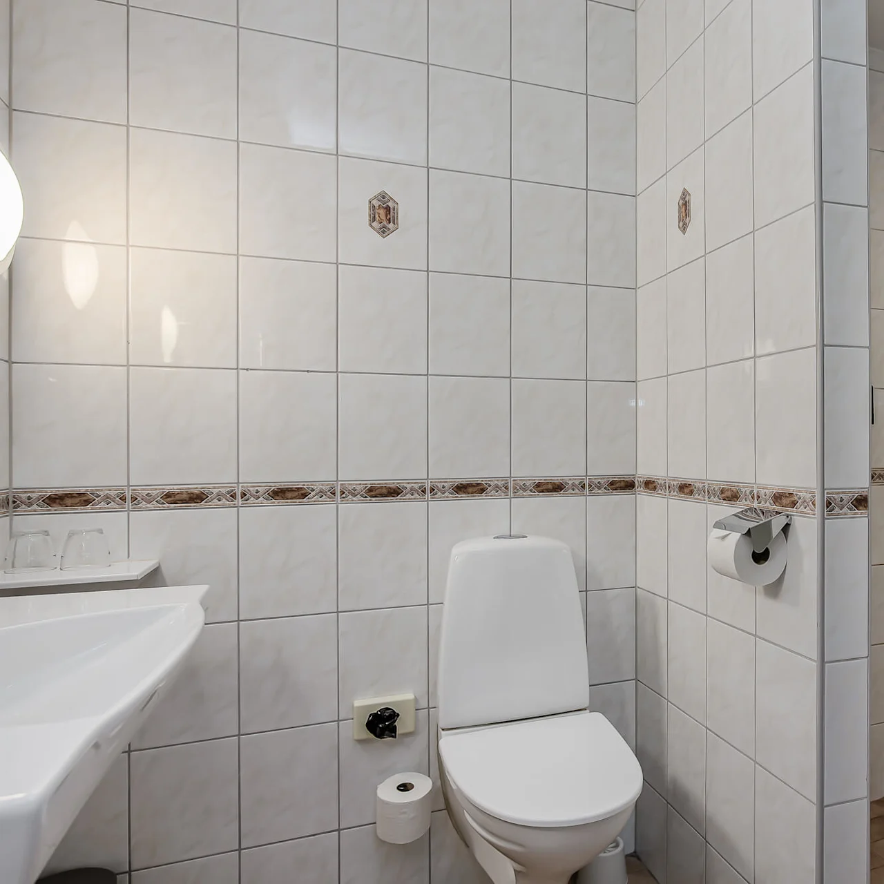 Valkoinen WC istuu pesualtaan vieressä laatoitetussa kylpyhuoneessa. Osittain avoin suihkutila näkyy, ja WC-paperi roikkuu seinällä.