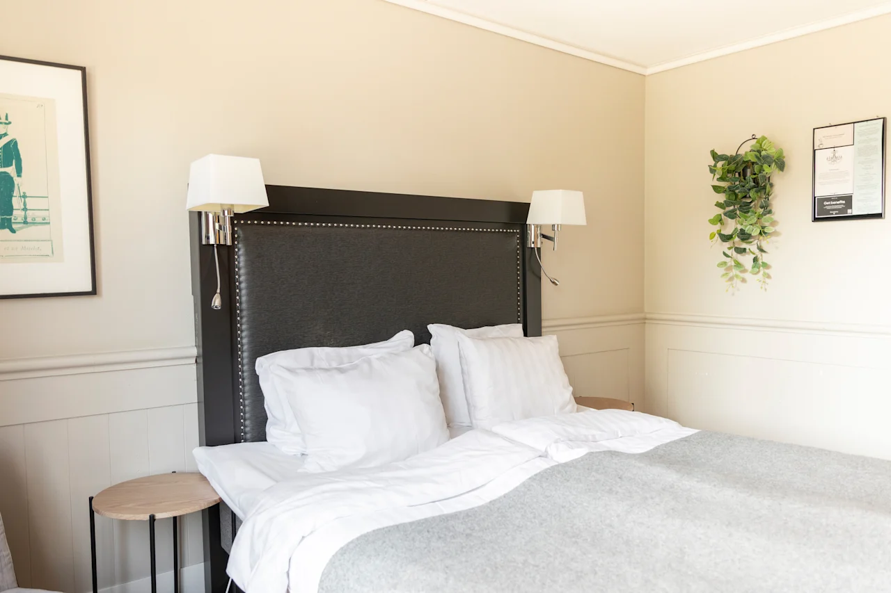 Ett mysigt sovrum med en prydligt bäddad säng, med en mörk sänggavel, vita sängkläder och vägglampor, vilket antyder komfort och avkoppling.