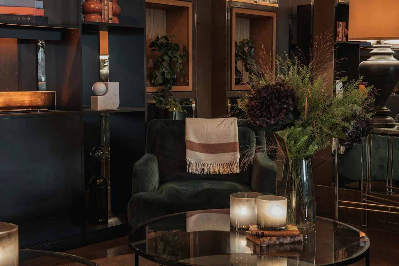 En mysig hotellounge med en grön fåtölj, ett soffbord med tända ljus och blommor, samt dekorativa hyllor. Home Hotel Grand, Helsingborg.