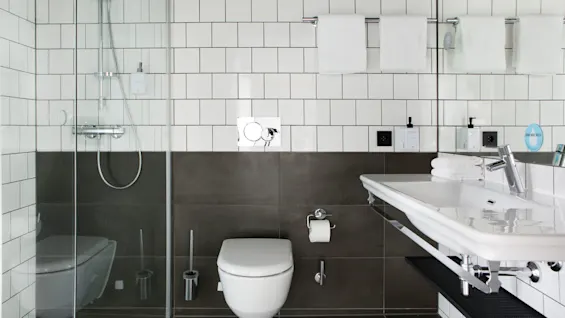Duschar med glasdörrar, ovanför en toalett, bredvid ett handfat, omgiven av vita och grå plattor. Handdukar hänger på ett ställ. En skylt lyder, "How much water?"