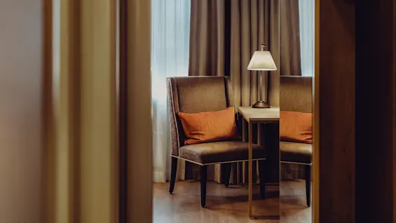 A cozy armchair with a cushion beside a lamp on an end table, located in a warmly lit room with curtains.