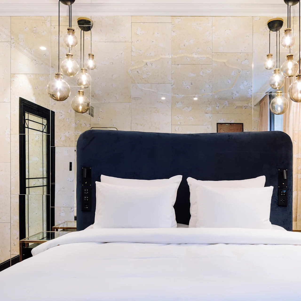 A bed with white linens and a dark headboard is centered under hanging globe lights in a modern, mirrored room with beige walls and a curtain.