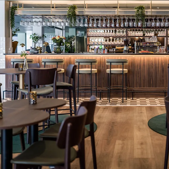 En bardisk med krakker omgitt av trebord og stoler. Dekor inkluderer diverse glass og grønne planter, noe som skaper en koselig, moderne atmosfære. Ingen tekst er til stede.