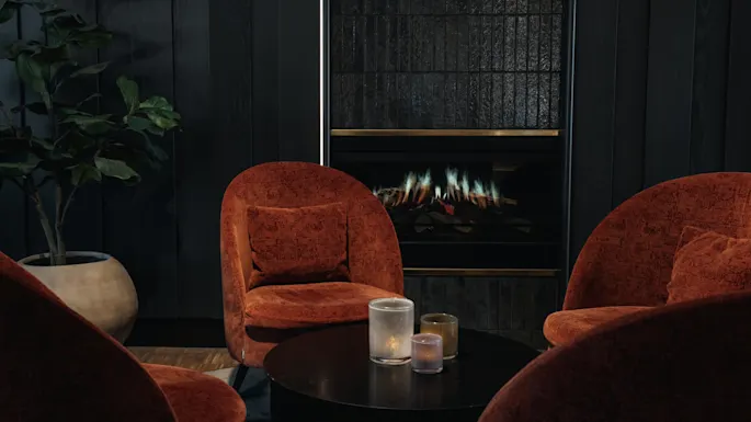 Et koselig salongområde med rustfargede lenestoler rundt et salongbord med stearinlys, mot en mørk vegg med peis.