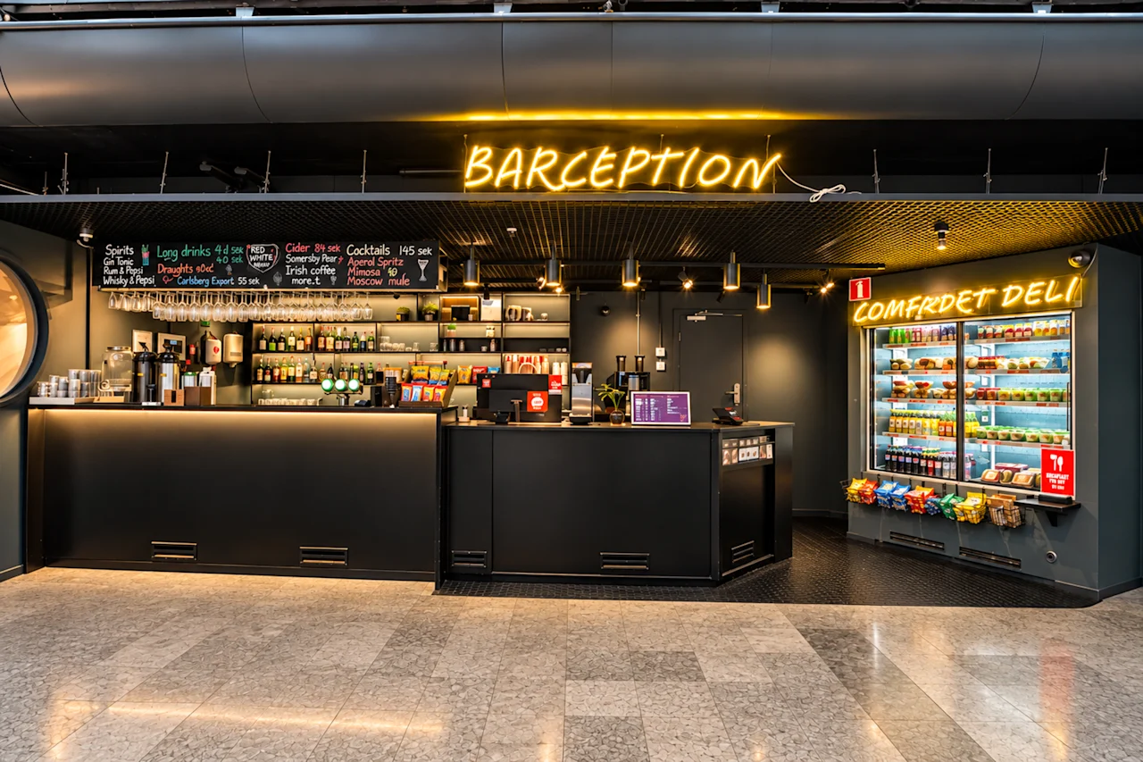 Modern hotel reception and bar with neon signs reading Barception and a well-stocked deli next to it.