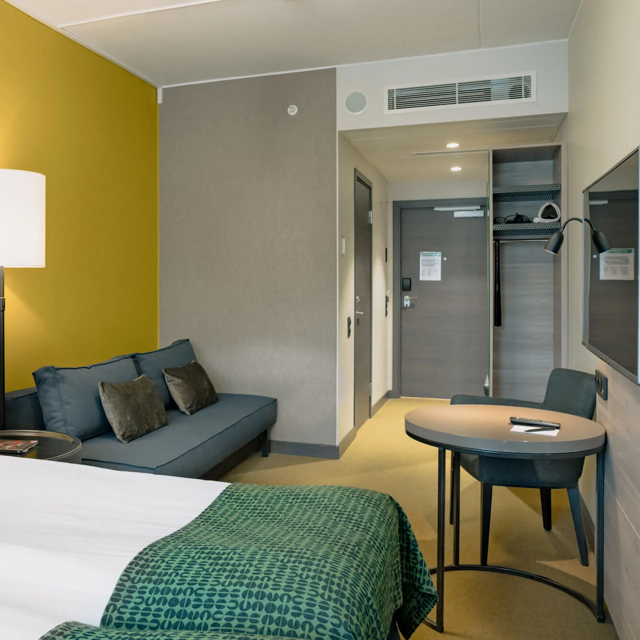 Ett hotellrum med en säng klädd i vitt och grönt linne, bredvid en blå soffa och ett bord med en stol, är upplyst av vägglampor mot en gul vägg.