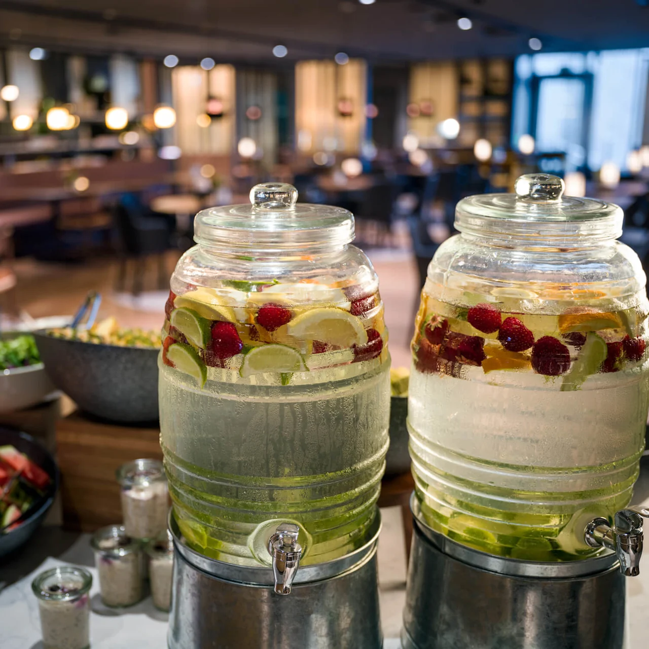 Glassbeholdere fylt med fruktinfusert vann, plassert på en buffébord med salat og oppskåret vannmelon, i et varmt, mykt opplyst spiseområde.