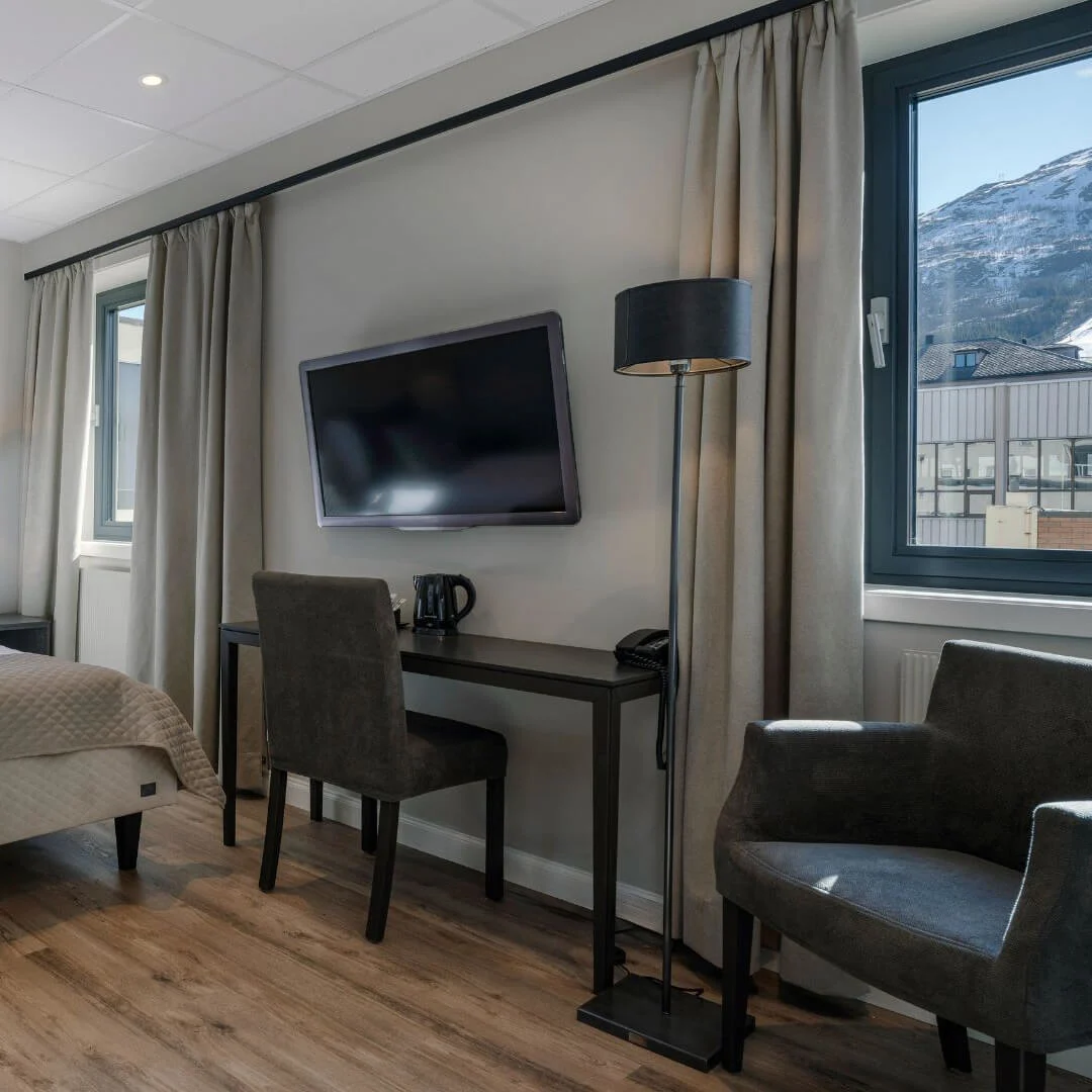 Sengen står sirligt redt, flankeret af to lamper, i et moderne hotelværelse. Et skrivebord har en kedel og kop under et vægmonteret TV, med bjerge synlige gennem vinduet.