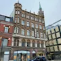 En fleretagers murstenshotelbygning med et grønt spir, med et "HOTELL"-skilt, står ved gaden, hvor folk går, og butikker, inklusive "Mulberry," kan ses nedenfor.