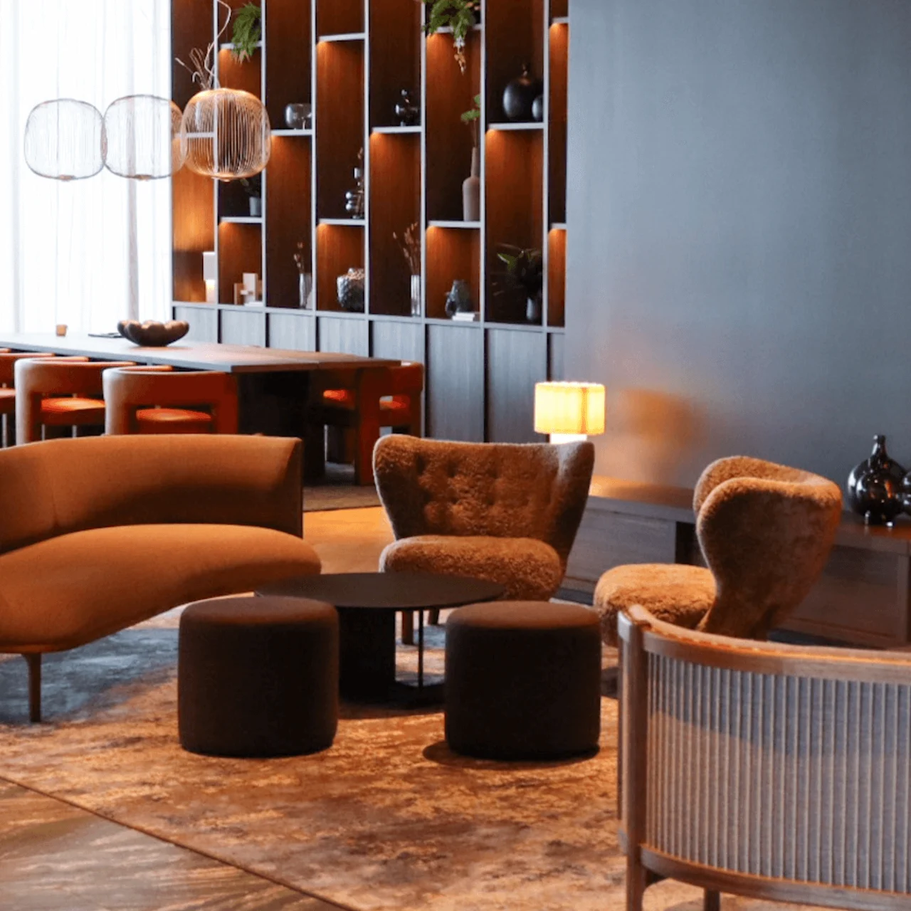 Modern chairs and tables surround a fireplace emitting a warm glow in a chic, open room with a decorative wall unit displaying vases and plants.