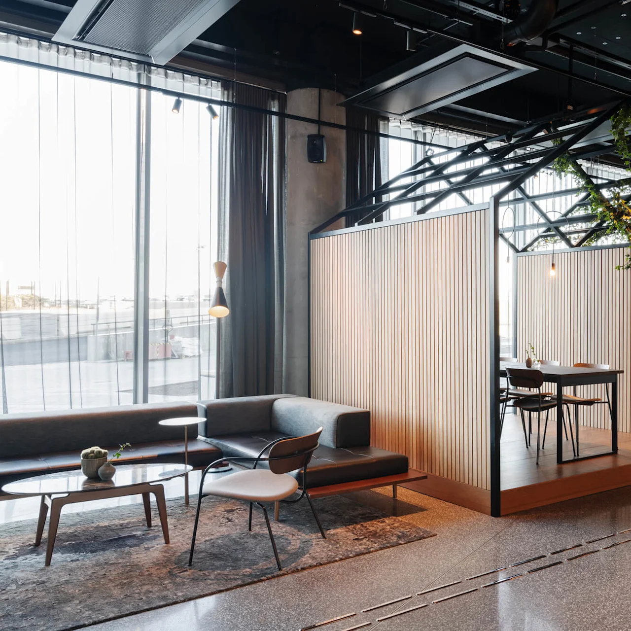 Cozy workspace booths with wooden walls feature tables and chairs, surrounded by a modern lounge with sofa and chairs. Overhead, greenery adds a natural touch.