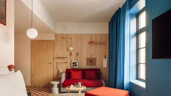 A vibrant hotel room with a red sofa, a wooden pegboard wall displaying a bag with NO BO, and a window with blue curtains.
