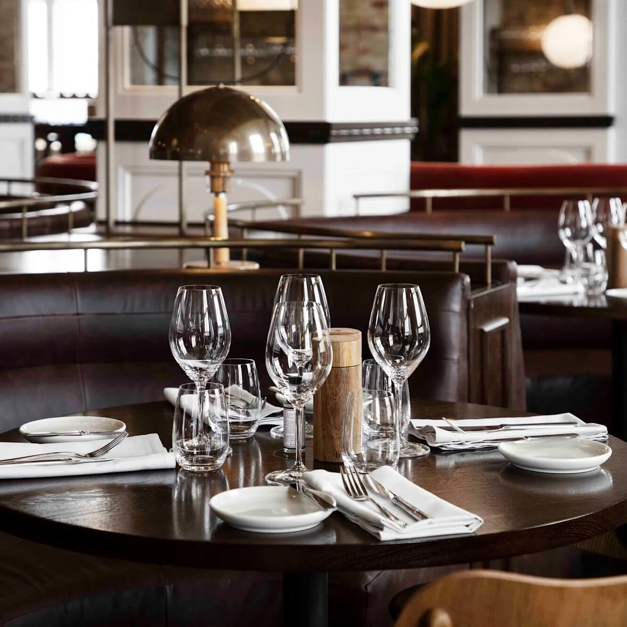 A round table set with wine glasses and napkins is surrounded by leather seating. The elegant restaurant interior includes cushioned booths, wooden chairs, and soft lighting.