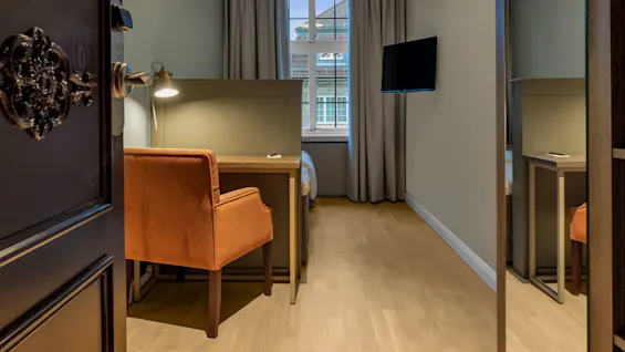 Ett hotellrum sedd från entrén: orangefåtölj, skrivbord, säng, tv och gardiner, med trägolv och dämpade färger.