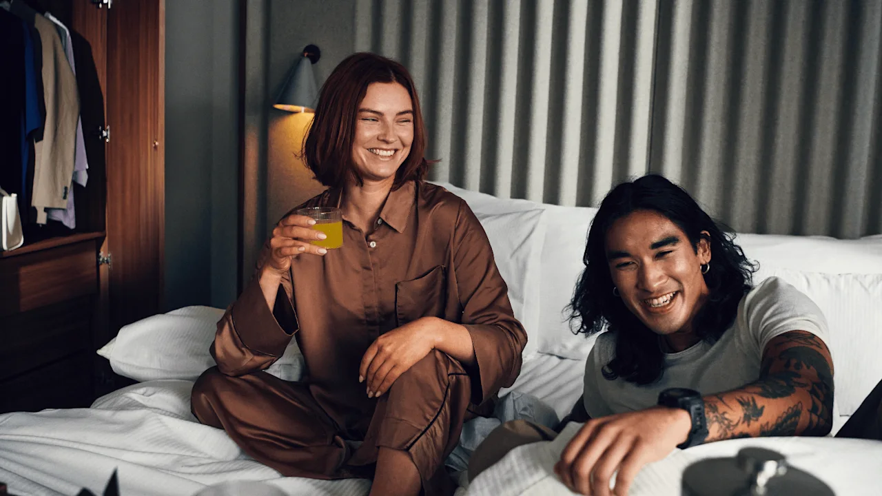 A person in silky pajamas holds a glass, smiling beside another person with tattoos, lounging on a bed in a warmly lit room with clothes visible in an open wardrobe.