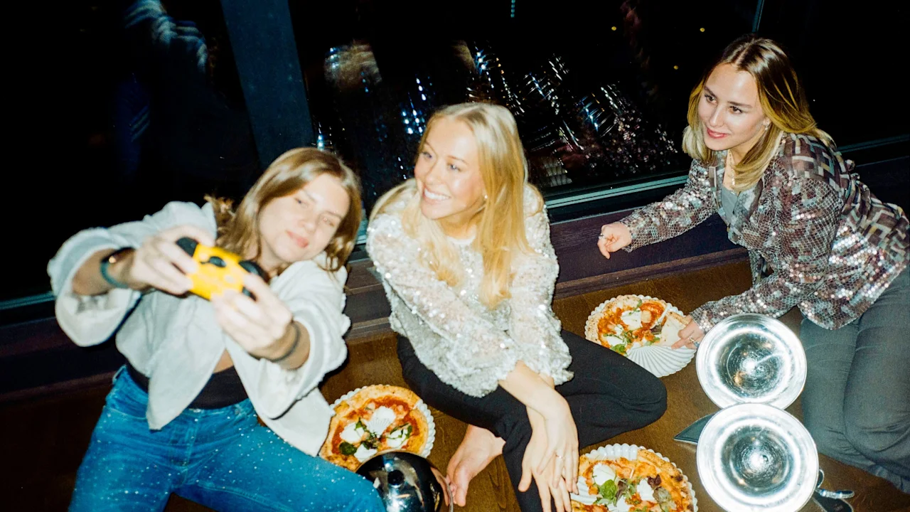 Venner samlet, deler pizzaer og fanger minner med et kamera, mot en bakgrunn av bylys om natten.