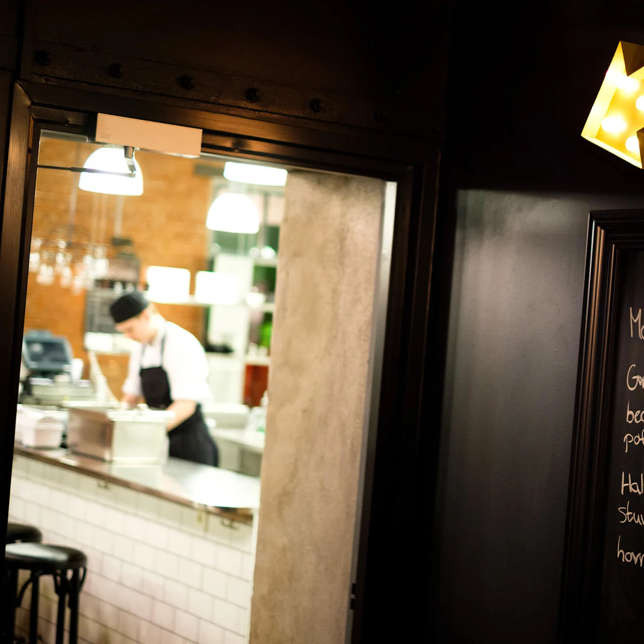 A chef works in a warmly lit kitchen, visible through a window; an arrow-shaped light points to a chalkboard menu.