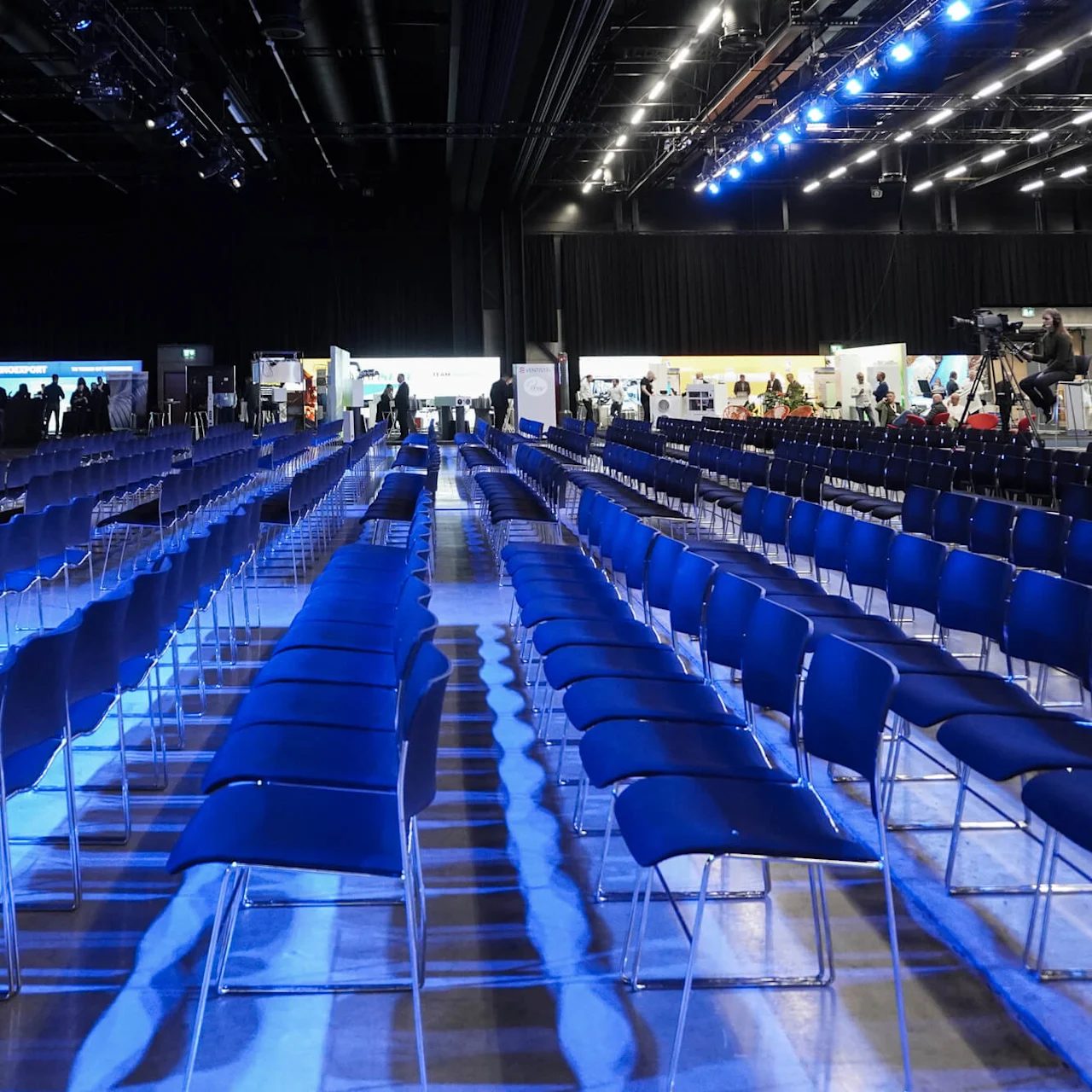 Rader av tomma blå stolar är upplysta av blå överljus i ett stort auditorium. Utställningsbås och personal syns i bakgrunden.