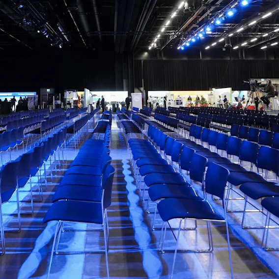 Rows of empty blue chairs are illuminated by overhead blue lights in a large auditorium. Exhibition booths and personnel are visible in the background. 