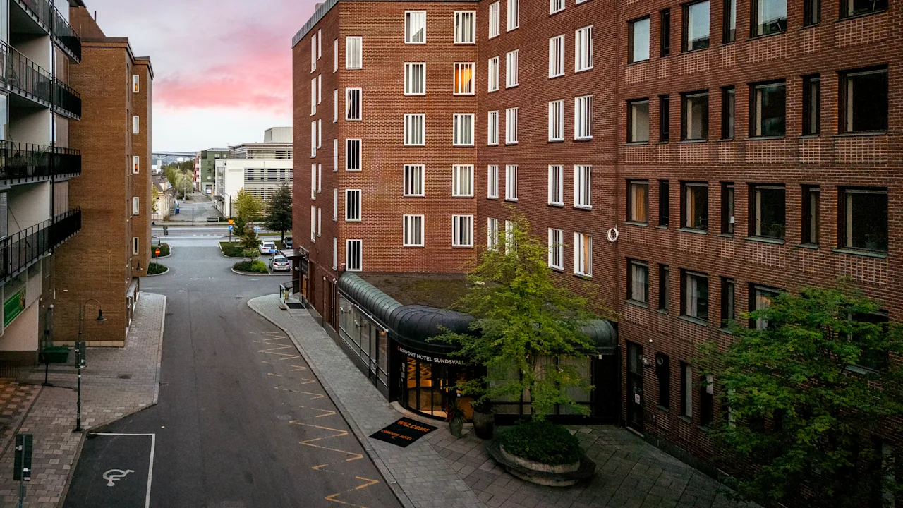 En gata kantad av höga tegelbyggnader vid solnedgång, med ett litet torg med träd och en byggnadsentré med tak som leder till en bredare väg längre bort.