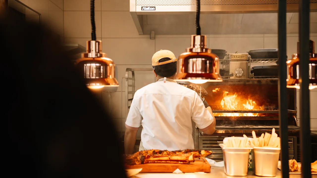 En kok tilbereder mad over åben ild i køkkenet på KG Hotel under varme lamper.