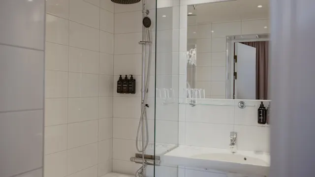 Duschhuvud hänger över ett badkar med glasvägg, omgivet av vita kakelplattor. Tvålpumpar är monterade på väggen. En handfat med spegel syns i närheten.