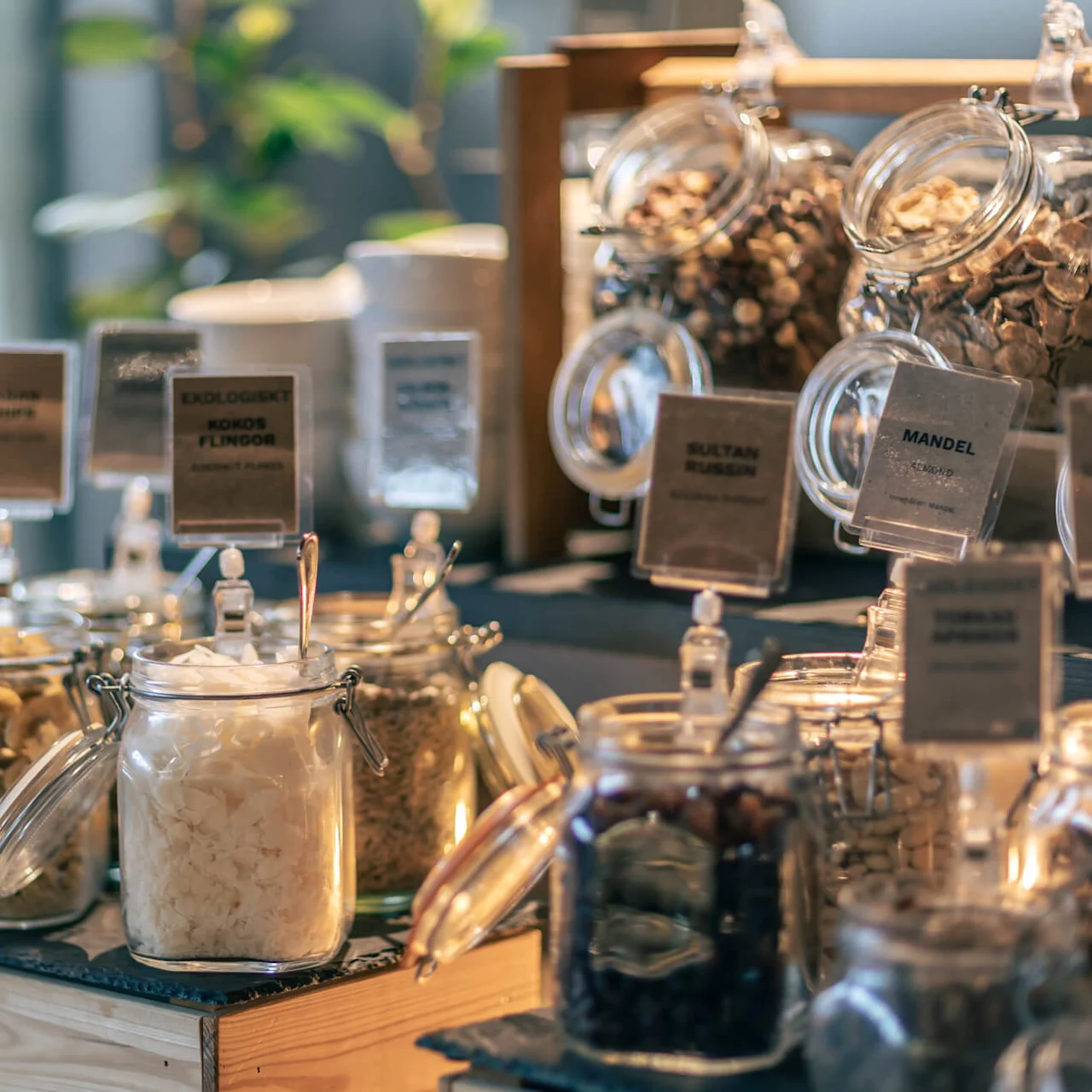 Jars filled with various dried foods, including labeled 'Ekologiskt Kokosflingor,' 'Sultan Russin,' and 'Mandel,' displayed on a wooden counter with spoons inside, in a bright setting.