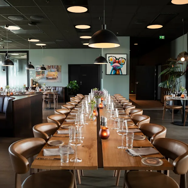 Et moderne restaurantinteriør med et langt spisebord dækket til en stor forsamling, omgivet af mindre borde.