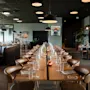 Et moderne restaurantinteriør med et langt spisebord dekket for en stor samling, omgitt av mindre bord.