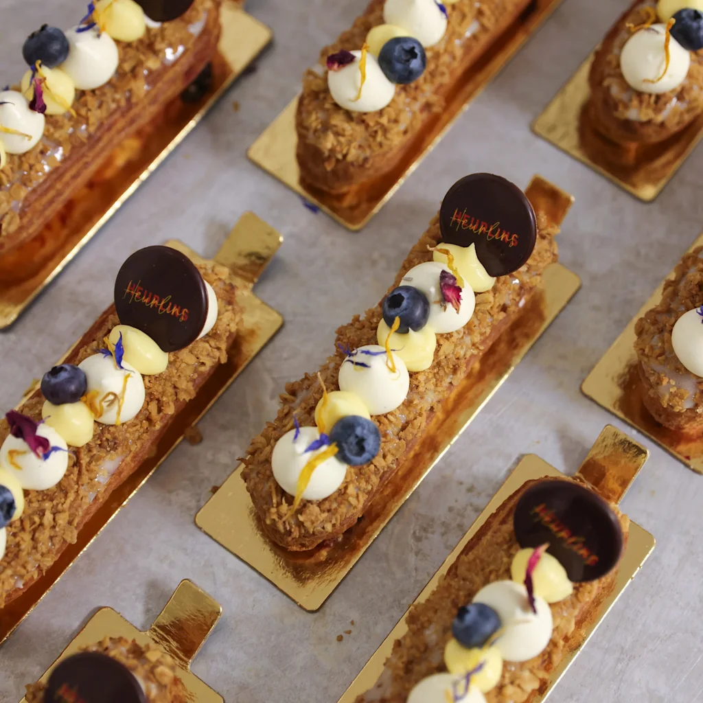 Rader av eleganta bakverk på gyllene brickor, toppade med grädde, blåbär och en chokladskiva med texten Heurlins.