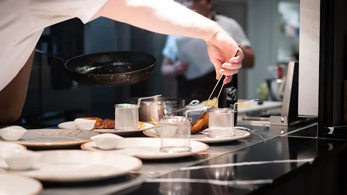 A chef arranges food with tweezers on a plate in a professional kitchen, surrounded by cooking utensils and a frying pan.