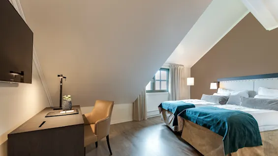Ett prydligt hotellrum har ett skrivbord med lampa och stol, bredvid en bäddad säng med teal-färgat täcke, under ett sluttande tak. Ett fönster släpper in naturligt ljus.