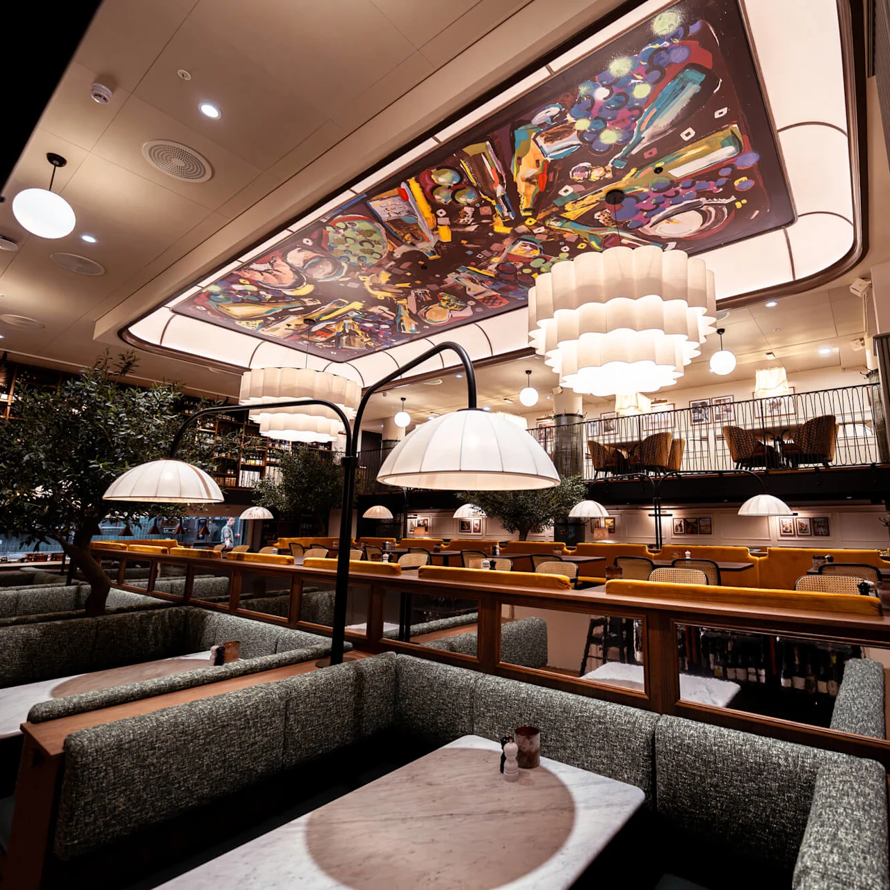 Ceiling art of vibrant colors decorates a stylish restaurant with modern lighting, plush booths, and wooden elements. The upper level features additional seating with woven chairs.