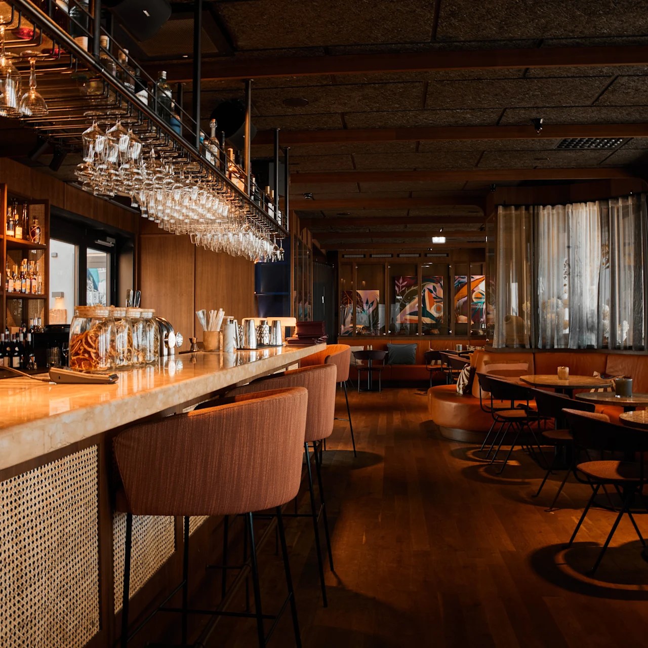Elegant bar med hängande glas över en polerad disk, med höga stolar och mysiga sittplatser i ett varmt belyst rum. Hyllor fyllda med flaskor ger en sofistikerad atmosfär.