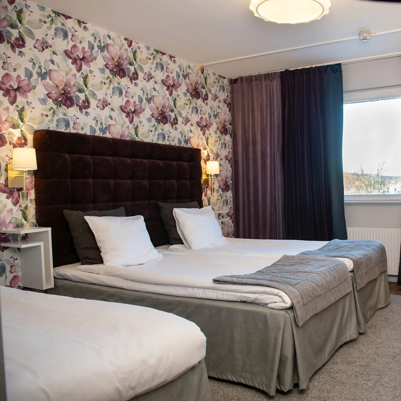 A hotel room features two white-covered beds against a floral wallpaper, with a dark headboard. Nearby, a desk with a lamp and chair sits beneath a wall-mounted TV.