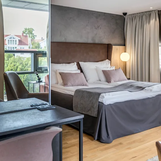 En pent og velstelt seng med puter og en brun hodegjerde står i et moderne rom med myk belysning og store vinduer, med et skrivebord og skinnstoler i nærheten.