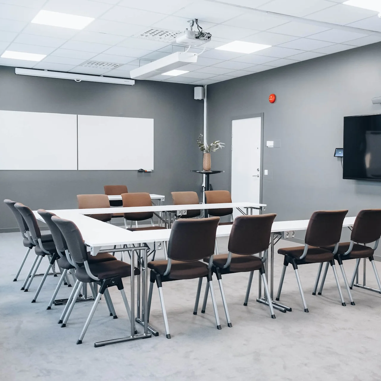 Conference room with arranged chairs and tables facing a large screen on the wall. Whiteboards and a potted plant decorate the gray interior, creating a professional meeting environment.