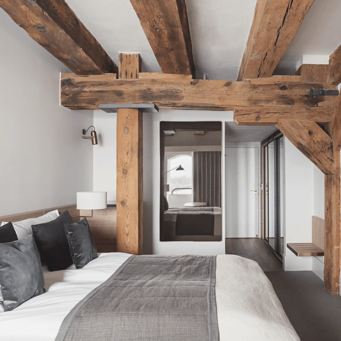 A bed with gray bedding is positioned under wooden beams in a modern room, facing a wall-mounted TV. A mirror reflects a windowed view.