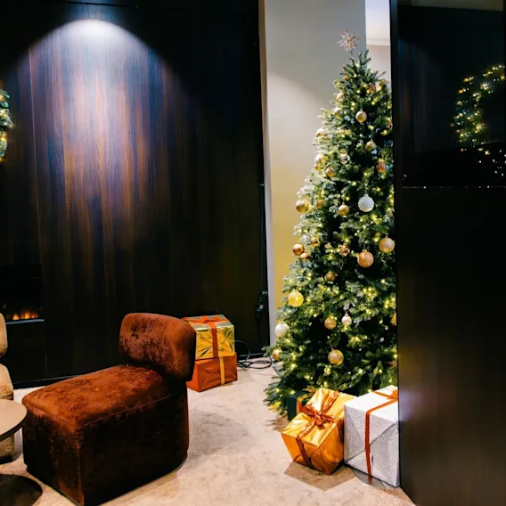 En koselig hotellounge julepyntet med et belyst juletre, gavedekorasjoner på gulvet, fløyelsstoler og en lysende krans.