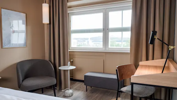 A cozy room features a bed, chair, and desk with a lamp. A large window lets in light, with curtains flanking it. A small round table sits nearby. No text.