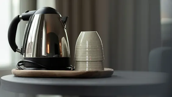 A shiny electric kettle and two stacked ceramic cups on a wooden tray, ready for a hot drink in a cozy room.