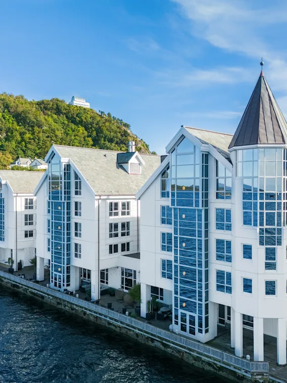 En moderne hvit bygning med store glassvinduer og et tårn, ved vannet med en grønn ås i bakgrunnen.