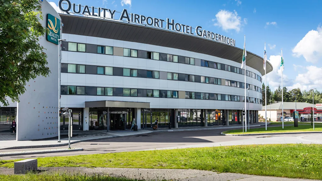 En moderne, buet hotellbygning merket "Quality Airport Hotel Gardermoen" med en klatrevegg, omgitt av trær og flagg under en klar blå himmel. Tekst inkluderer "Quality Hotel."