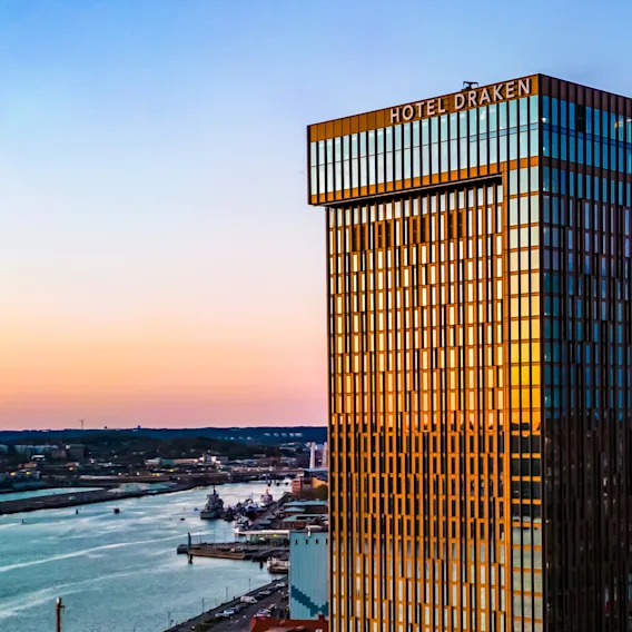 A tall, modern hotel building labeled "HOTEL DRAKEN" reflects sunlight, standing beside a river at sunset, surrounded by cityscape elements including buildings and a bridge.