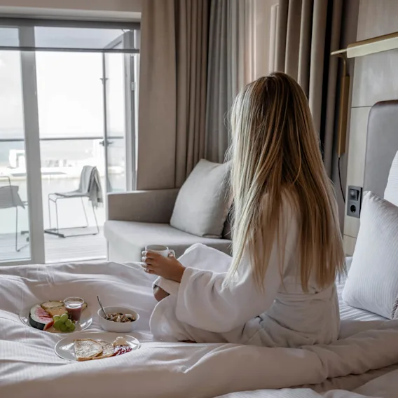 En kvinna i vit morgonrock sitter på en säng och håller i en mugg, med frukosttallrikar i närheten. Hon tittar ut genom ett fönster mot vatten och byggnader.