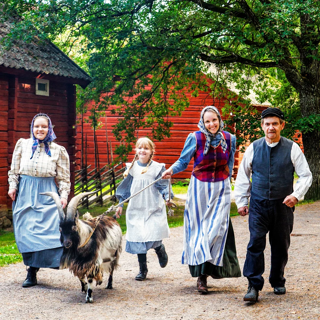People in traditional attire walk a goat on a leash at Vallby friluftsmuseum, with red wooden buildings in the background.