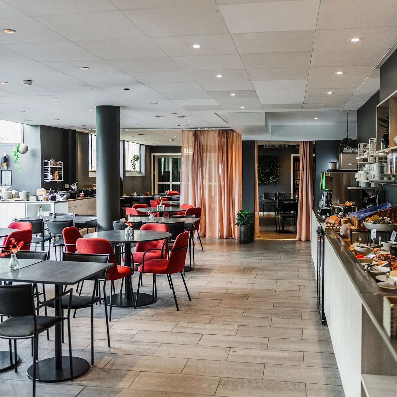 Cafeteria med moderna bord och röda stolar, med en buffé till höger med olika maträtter. Utrymmet har mjuk belysning och enkel inredning.