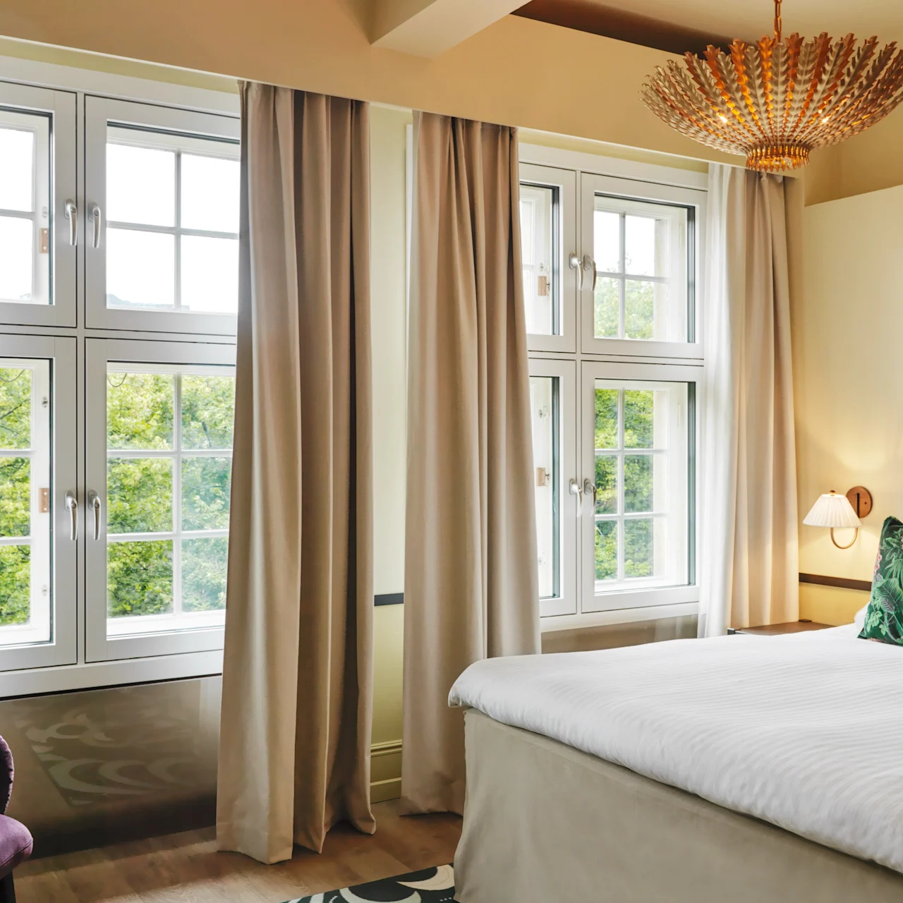Bed stands neatly in a bright room with large windows and beige curtains, under a stylish golden chandelier, complemented by a teal headboard and floral cushions.