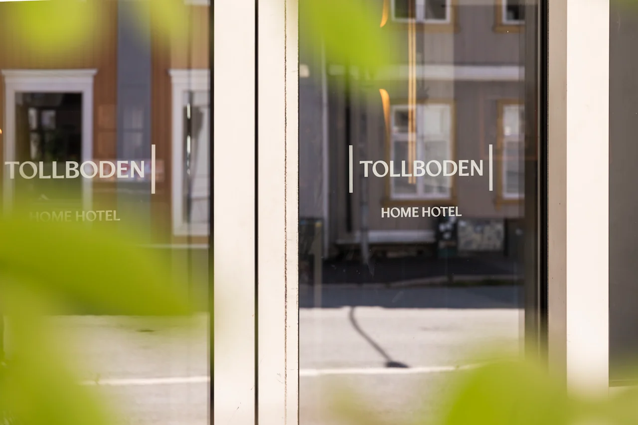 Glasdörrar visar texten "TOLLBODEN HOME HOTEL" och reflekterar en gata med byggnader. Gröna löv ramar delvis in vyen, vilket indikerar en utomhusmiljö.